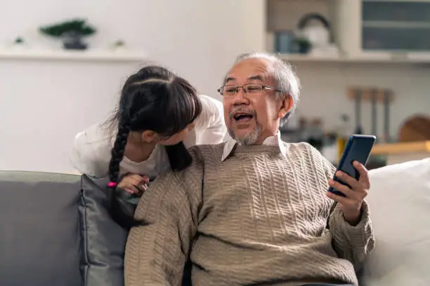 Happy retirement elderly man sitting on sofa at living room with granddaughter using mobile phone together. Multigenerational family with technology concept.