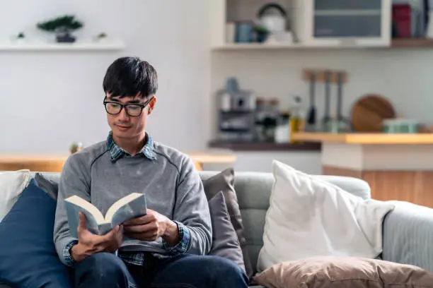 Happy asian young adult man sitting on sofa at living room reading fiction book. Domestic life relax at home.