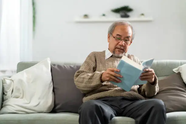 Happy asian retirement elderly man sitting on sofa at living room reading fiction book. Domestic senior life at home.