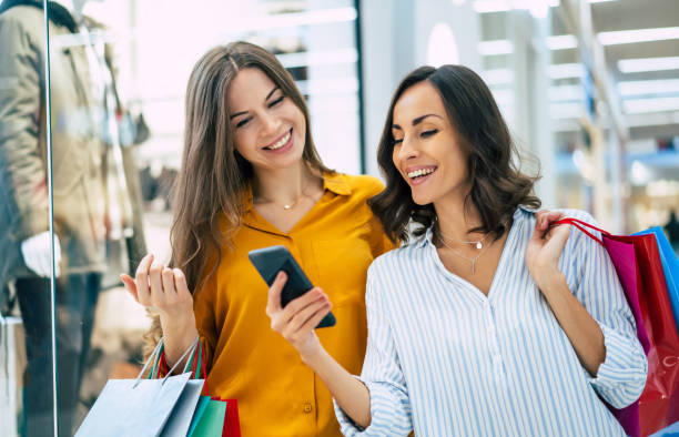 lindas amigas felizes e animadas com sacos de papel e telefone inteligente estão andando pelo shopping - shopping women internet credit card - fotografias e filmes do acervo