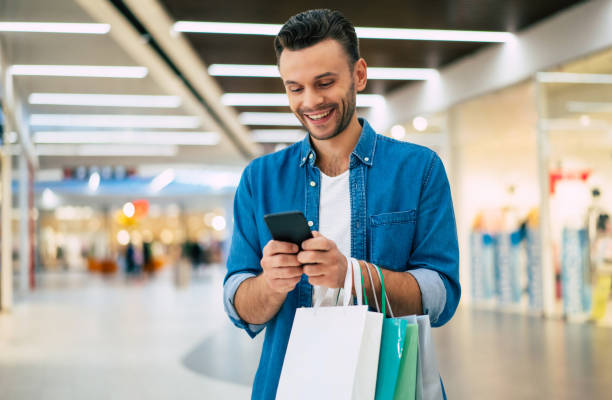 handsome smiling young stylish bearded man with shopping bags is using smart phone while walking in the mall on black friday - telephone indoors retail shopping mall imagens e fotografias de stock