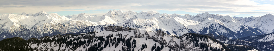 Winter snow mountain ranges and woodland. Beautiful snowy winter landscape panorama in Alps with orange sunset sunlight. View from Riedberger Horn to Grasgehren Ski Resort and Allgauer Alps. Allgau, Bavaria, Germany.
