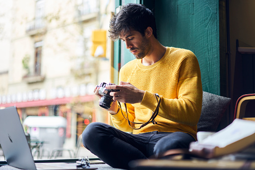 Thoughtful ethnic male photographer in casual wear sitting on comfortable windowsill in cozy cafe and browsing camera while enjoying time