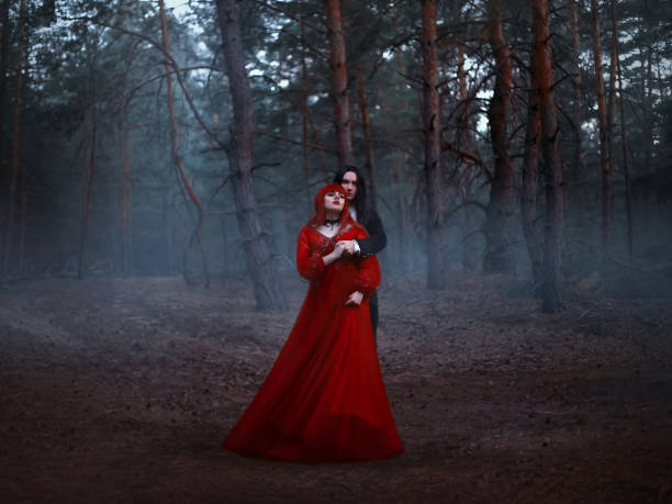 blurred silhouette of a gothic couple in motion. cinema noise and grain added, old film style. a vampire man in a black tailcoat with long hair embraces a woman in red medieval dress. deep dark forest - long hair red hair women men imagens e fotografias de stock
