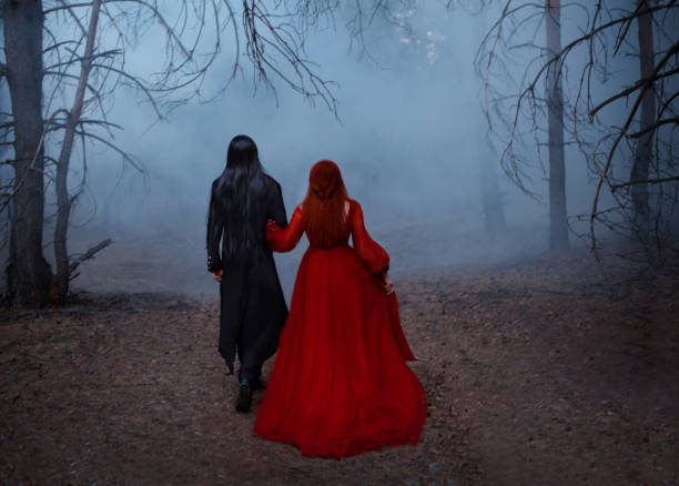 cinema noise and grain added, old film style. blurred silhouette of a gothic couple in motion. a vampire man in a black tailcoat walks with a woman in red medieval long vintage dress. deep dark forest - long hair red hair women men imagens e fotografias de stock