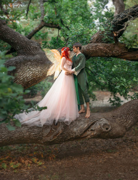 jeune couple dans l’amour. un homme et une femme s’étreignant sur un arbre énorme. sur le sujet de mariage créatif photographie fantastique lumineux. femme de fée dans la longue robe rose avec les ailes dorées lumineuses. un homme dans un costume  - fairy forest women magic photos et images de collection