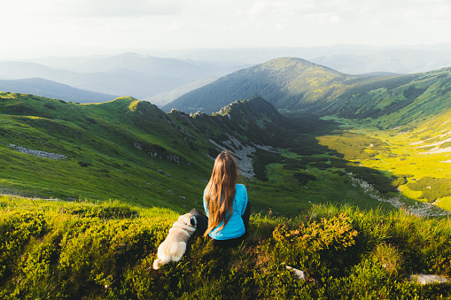 Woman with long hair and her cute small dog - pug breed enjoying the scenic sunset above the mountain peaks sitting on the green fluffy meadow
