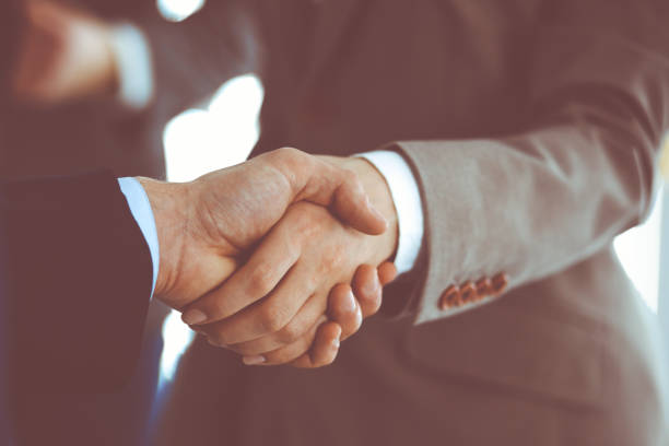 istock Business people shaking hands while standing with colleagues after meeting or negotiation, close-up. Group of unknown businessmen and women in modern office. Teamwork, partnership and handshake concept, toned picture 1289023505