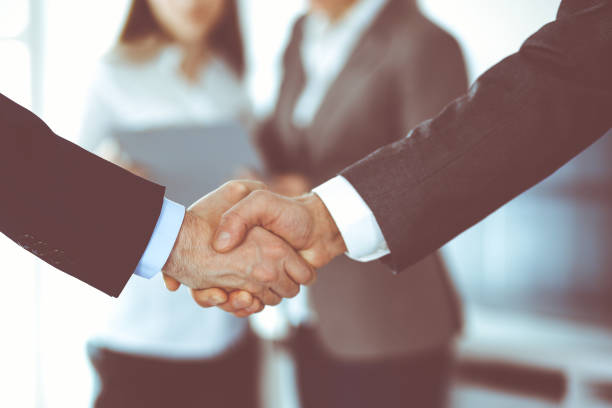 istock Business people shaking hands while standing with colleagues after meeting or negotiation, close-up. Group of unknown businessmen and women in modern office. Teamwork, partnership and handshake concept, toned picture 1289023542