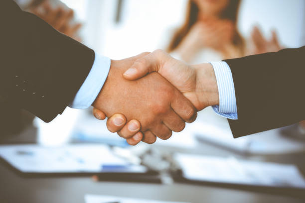 istock Business people shaking hands at meeting while theirs colleagues clapping and applauding. Group of unknown businessmen and women in modern white office. Success teamwork, partnership and handshake concept 1289025333