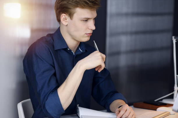 istock Young blond businessman thinking about strategy at his working place with computer in a darkened office, glare of light on the background 1289027070