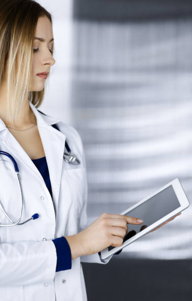 istock Young professional woman-doctor is using a tablet computer, while standing in a clinic. Portrait of beautiful female physician at work. Perfect medical service in hospital. Medicine concept 1289030131