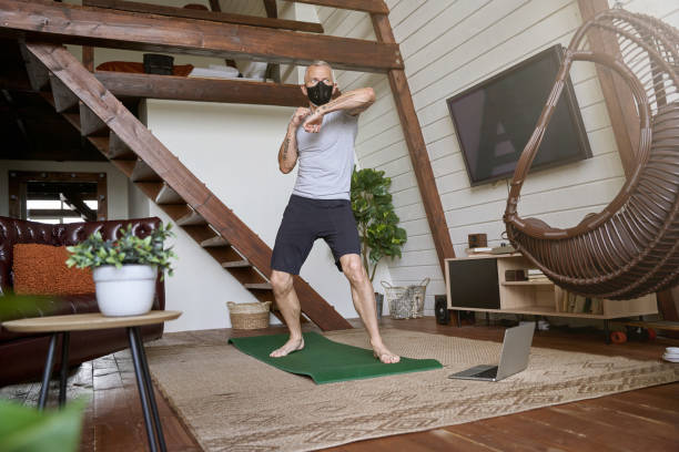 istock Active middle aged man wearing face protective mask standing in boxing stance while exercising at home, watching online workout on laptop. Sport during covid 19 1289030739