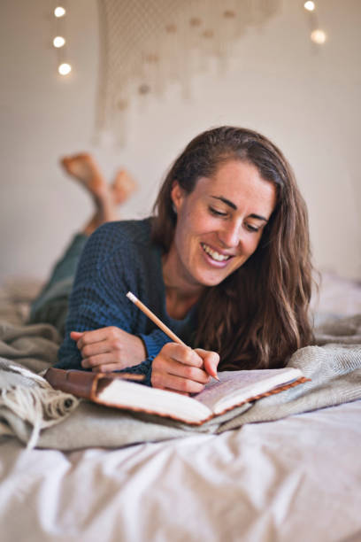 Woman lying on her bed writing in her notebook Woman lying on her bed writing in her leatherbound notebook. bullet journal photos stock pictures, royalty-free photos & images