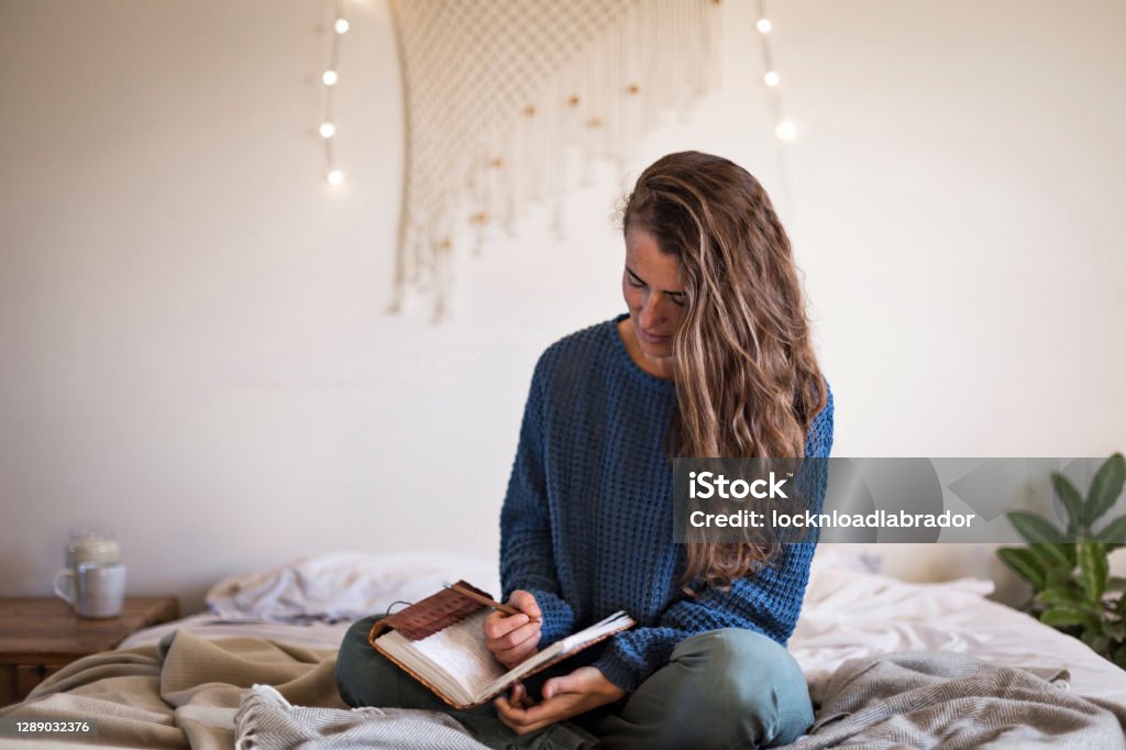 Woman sat on bed writing in her journal Woman in blue sweater sat on her bed writing in her leatherbound journal Bullet Journal Stock Photo