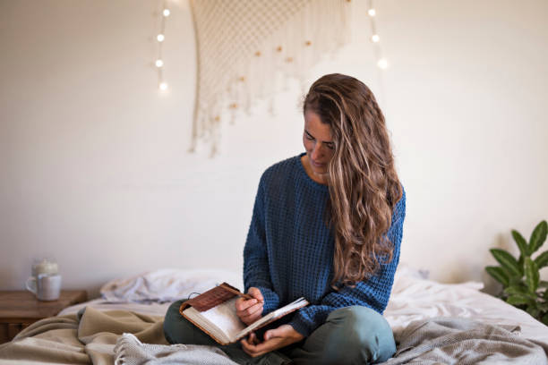 Woman sat on bed writing in her journal Woman in blue sweater sat on her bed writing in her leatherbound journal bullet journal photos stock pictures, royalty-free photos & images