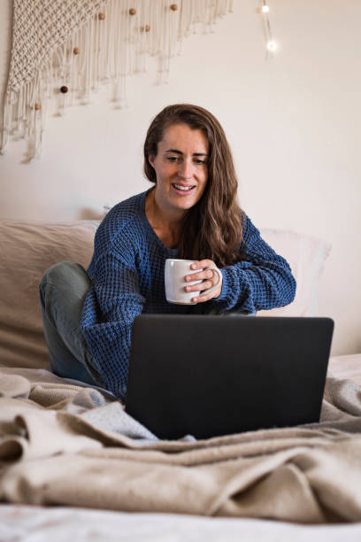 istock Woman sat on bed using laptop computer and drinking mug of tea 1289035512