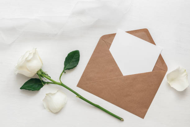 istock Wedding invitation. Blank card in brown envelope mock-up, white rose and silk ribbon. White wooden painted background. Top view, flat lay. 1289035561
