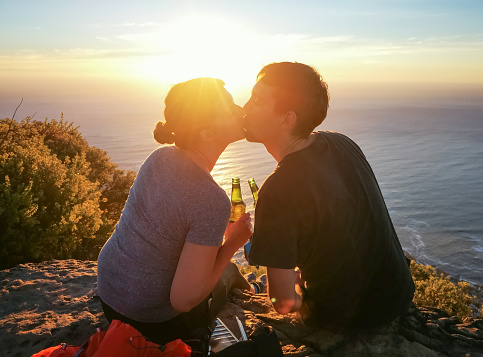 Young couple kissing with beer in front of elevated sunset ocean view, feeling a moment of pure happiness.