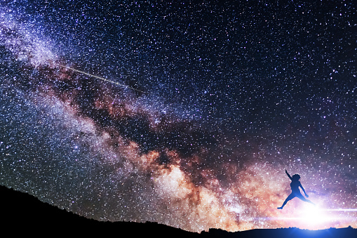Silhouette of a person jumping over the hill on the bright milky way galaxy. Beautiful strry sky. Astronomical background.