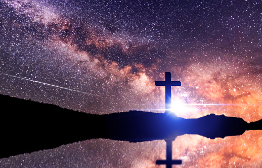 The cross silhouette on on the hill in night , behinde bright milky way galaxy and light rays.