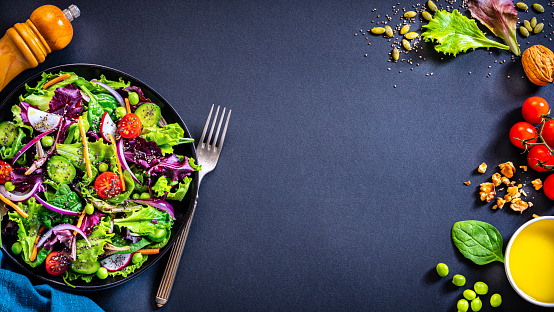 Vegan food: overhead view of a healthy fresh vegetables salad plate shot on dark background. Chia seeds, pumpkin seeds, walnut and lettuce leaves are around the salad plate. The composition is at the left of an horizontal frame leaving useful copy space for text and/or logo at the right. High resolution 35,5Mp studio digital capture taken with Sony A7rII and Sony FE 90mm f2.8 macro G OSS lens