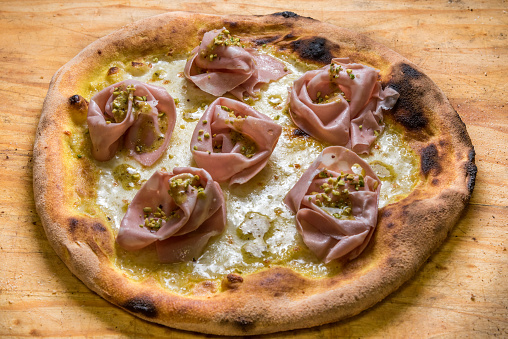 pizza with cheese, mortadella salami from Bologna and chopped pistachios, typical Italian food on rustic wooden cutting board