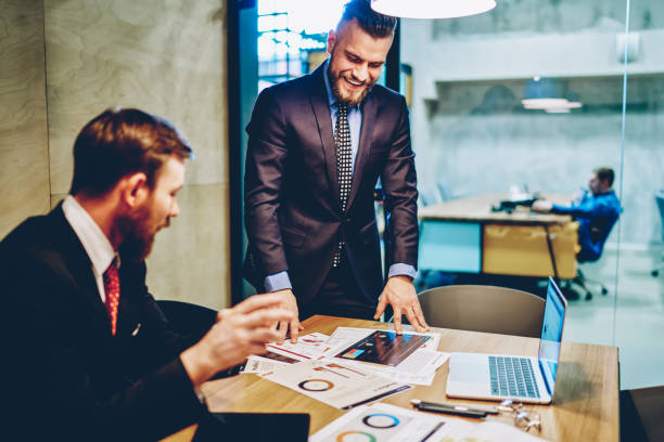 Cheerful male marketers analyzing financial statistics during productive collaboration at table desktop with laptop, successful businessmen enjoying friendly brainstorming for discuss documents