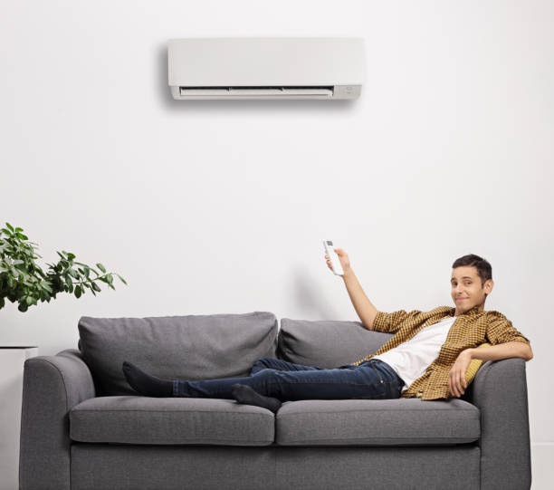 istock Young man sitting on a sofa and putting an air conditioning unit on with a remote control 1289073080