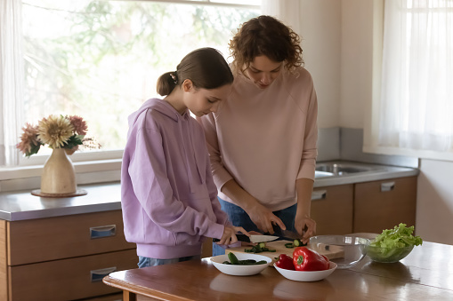 Loving Caucasian mother teach teenage daughter cooking healthy food at home kitchen. Caring mom and teen girl child prepare salad for dinner lunch, cut fresh organic bio vegetables. Diet concept.