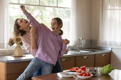 Happy Caucasian mom and teen 13s daughter have fun dancing signing while cooking together at home kitchen. Smiling mother and teenage girl child feel playful crazy preparing food in the morning.