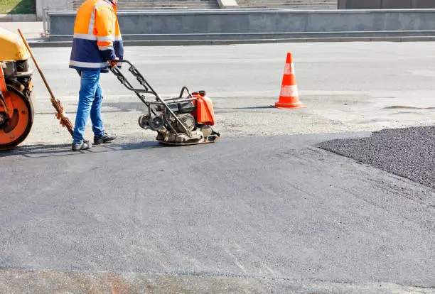 A road worker compacts asphalt on a fenced road section of the roadway with a petrol vibration compactor.