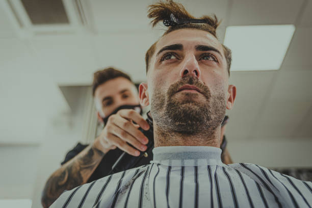 a young man with tattooed arms cuts a man's hair in a barbershop stock photo