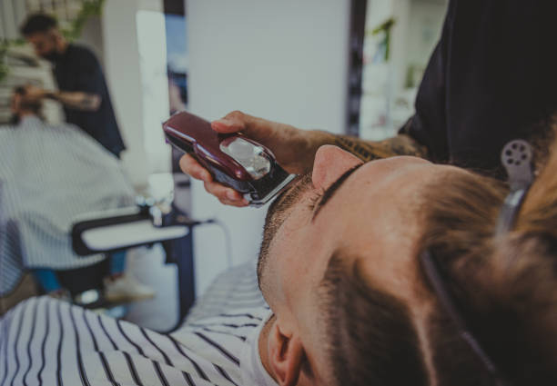 detail of a young man with tattooed arms cuts a man's hair in a barber shop stock photo