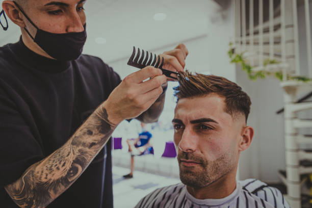 a young man with tattooed arms cuts a man's hair in a barbershop stock photo