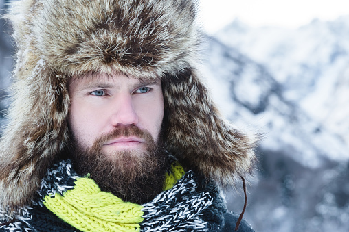 Man with a beard in scarf and a hat stands in the mountains in winter. Active recreation concept.