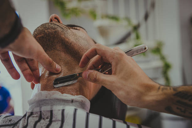 detail of a young man with tattooed arms cuts a man's hair in a barber shop stock photo