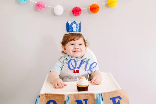 Cute adorable Caucasian baby boy in blue crown celebrating his first birthday at home. Child kid toddler sitting in high chair eating tasty cupcake dessert with word topper. Happy birthday concept.