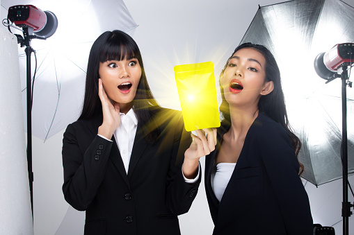 Two asian women shows green package which has empty green area for advertising, in studio with umbrella light background copy space