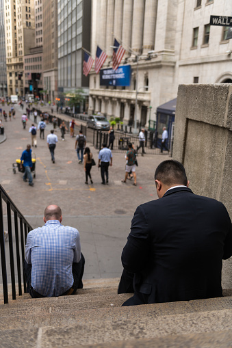 New York, United States - Jul 22,  2019 Two business men sitting on the steps with the Stock Exchange in the distance mid day.