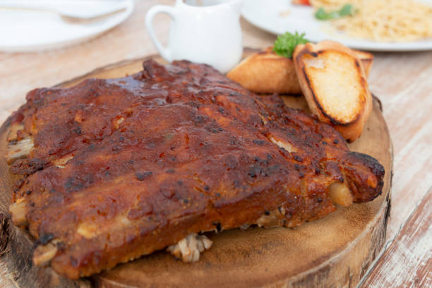 Close up view of barbecue ribs with and sauce on wooden table. Pork ribs on a wooden plate. Hot grilled spare ribs served with bread and fresh vegetables on an old vintage wooden board. vista de cerca de las costillas de barbacoa con y salsa en la mesa de madera. costillas de cerdo en un plato de madera. costillas de repuesto a la parrilla calientes servidas con pan y verduras frescas en una vieja tabla de madera vintage.