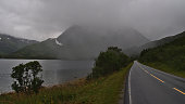 View of empty paved country road 85 at Gullesfjord, Hinnøya, Vesterålen in northern Norway on rainy day with green vegetation, fjord and majestic mountains disappearing in the low clouds.