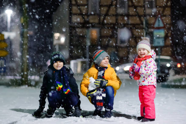 два маленьких мальчика и милый малыш девушка сидит вместе играть со снегом в зимнюю ночь. братья и сестры, братья и младшая сестра наслаждаю - little boys sled clothing slide стоковые фото и изображения