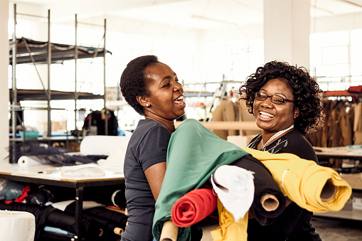 Two black African Zimbabwean woman smiling and laughing working together in a textile factory carrying many different colour rolls of fabric for processing