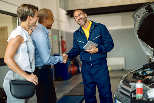 Happy auto repairman greeting a couple in a workshop. Men are shaking hands.