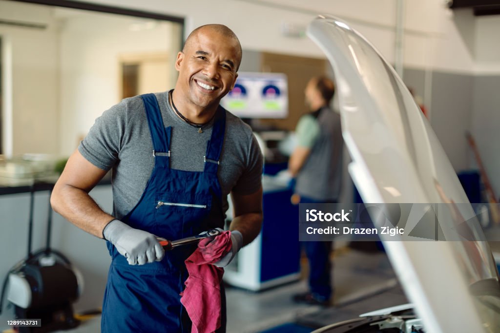 Happy black auto mechanic working at car workshop and looking at camera. Happy African American mechanic working in auto repair shop and looking at camera. Mechanic Stock Photo
