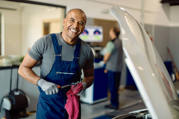 szczęśliwy czarny mechanik samochodowy pracujący w warsztacie samochodowym i patrzący na kamerę. - mechanic zdjęcia i obrazy z banku zdjęć