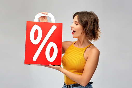 sale, consumerism and people concept - happy smiling young woman in mustard yellow top holding red shopping bag with percentage sign over grey background