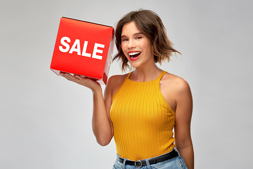 shopping and people concept - happy smiling young woman in mustard yellow top and jeans with sale sign posing over grey background