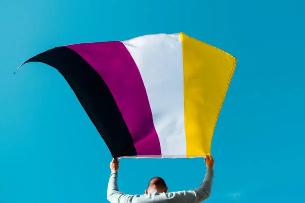 closeup of a young caucasian person, seen from behind, waving a non-binary pride flag on the air